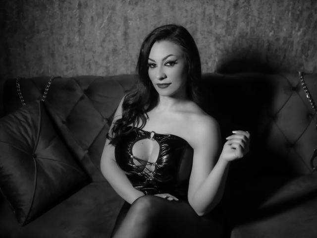 AmberCrost with long dark hair, wearing a black leather strapless dress with a cutout, sitting on a dark tufted sofa, holding a cigarette, black lipstick, sexy.