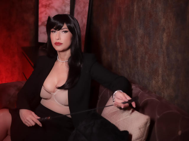 MissVonTease with black bob, red lipstick, black blazer, beige bra, showing tits, sitting on brown couch, holding remote, red light, silver necklace, serious expression.