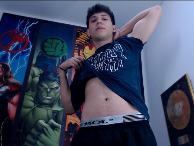 JonathanMoreau with short black hair, lifting a dark green shirt to reveal his flat, pale belly and black BOL.com underwear, standing in front of colorful superhero posters.