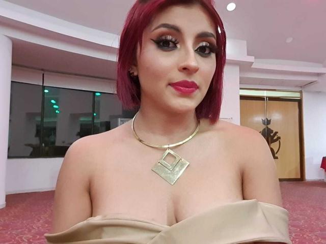 LizBonnet has red hair, heavy eyeliner, pink lipstick, gold necklace, and is wearing a strapless beige dress, showing her tits. She's smiling in a well-lit room.