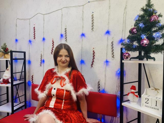 FreyaDak with long brown hair, smiling, wearing a red velvet Santa dress with white fur trim, sitting on a red bench, Christmas decorations, blue lights, small tree, and shelves.