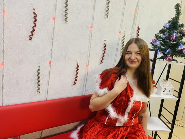 FreyaDak with long brown hair, smiling, wearing a red velvet Christmas dress with white fur trim, holding the dress, sitting on a red bench, next to a decorated tree.