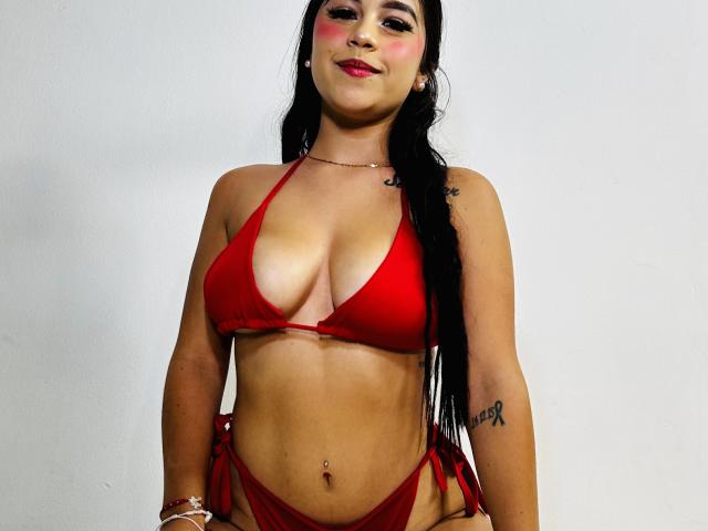 LucyCute with long black hair, red bikini, pink blush, smiling, showing her tits, tattoo on right arm, belly button piercing, white background, red string bikini bottom.
