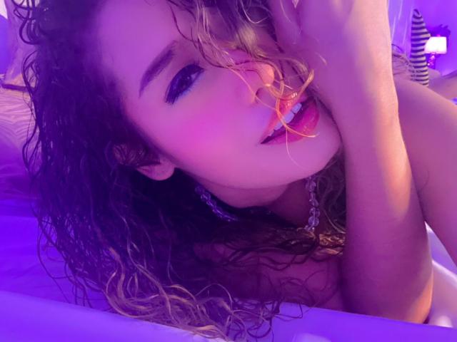 AkiraZanoli with curly blonde hair, purple eye makeup, and chain necklace, bites her finger, looking seductive, in purple-lit photo, showing her tits and pouty lips.