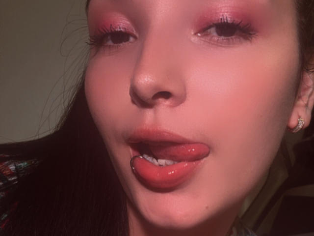 O rosto de SarahGood, com sombra rosa e lábios vermelhos brilhantes, com a língua ligeiramente para fora, mostrando uma pequena piercing de metal na parte inferior do lábio. Sua pele é clara e ela está usando brincos..