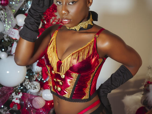 EbonyM mit dunkler Haut, trägt ein glänzendes, rot-goldenes, fransiges Korsett, schwarze Handschuhe und eine goldene Halskette, und steht vor einem geschmückten Weihnachtsbaum. Brüste sind sichtbar..