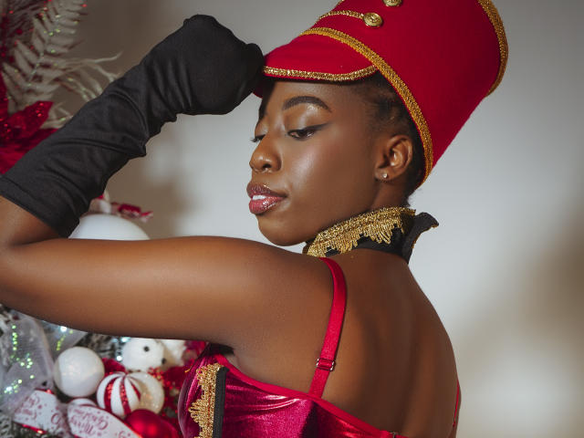 EbonyM mit dunkler Haut, die einen roten und goldenen Hut, schwarze Handschuhe und ein rotes Samtkleid trägt, zeigt ihren Rücken und dreht ihren Kopf leicht, wobei ihre glänzlichen Lippen und die goldene Halskette zu sehen sind..