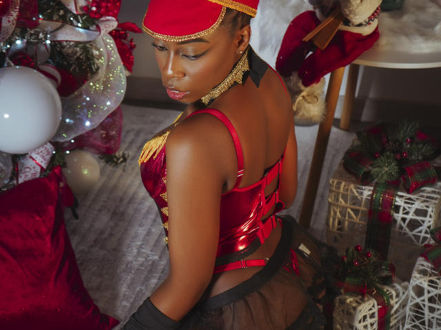 EbonyM, con la pelle scura, indossa una parte superiore in velluto rosso e una catena dorata, una gonna nera trasparente, un copricapo rosso, guarda indietro, mostrando il sedere, vicino a un albero di Natale con decorazioni..