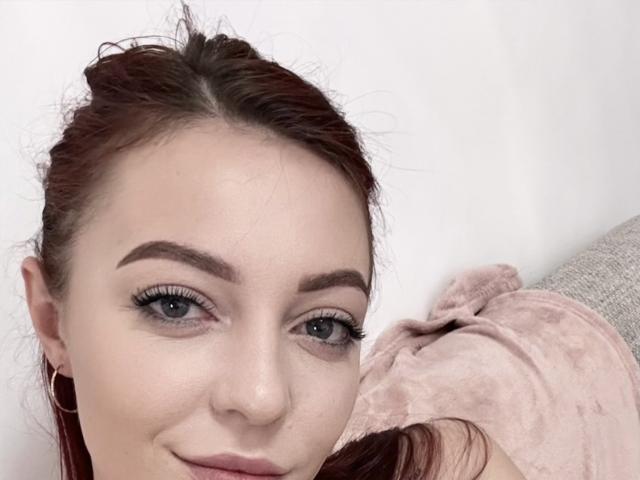 JustKessie with red hair, blue eyes, and thick eyebrows, wearing hoop earrings, smiling slightly, showing off her pretty face and subtle makeup. She's lying on a pink blanket.