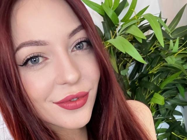 JustKessie has red hair, blue eyes, and red lipstick, with a subtle smile and winged eyeliner. Her tits are not visible. Green plant in the background.
