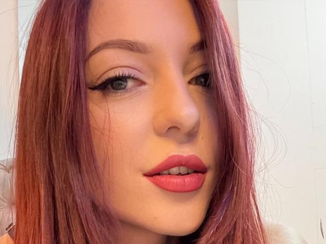 JustKessie has red hair, green eyes, and pink lipstick, with neatly lined eyes and slightly parted pink lips, looking slightly to the side in a selfie.