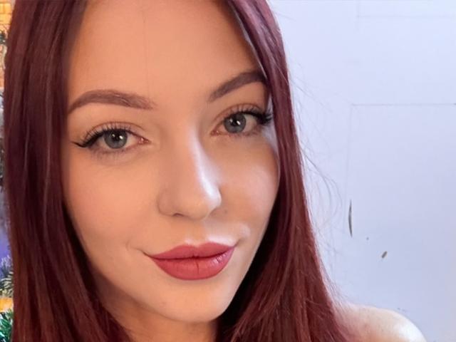 JustKessie has red hair, blue eyes, and is smiling with pink lipstick. Her eyelashes are long and she's looking at the camera. She has a cute, attractive face.