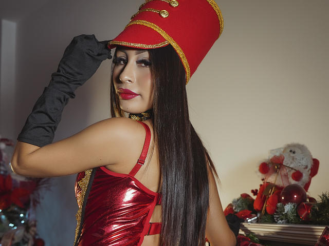 MiaFisher with long brown hair, red lipstick, and a shiny red dress, wearing black gloves and a red hat with gold trim, looking back, showing her ass.