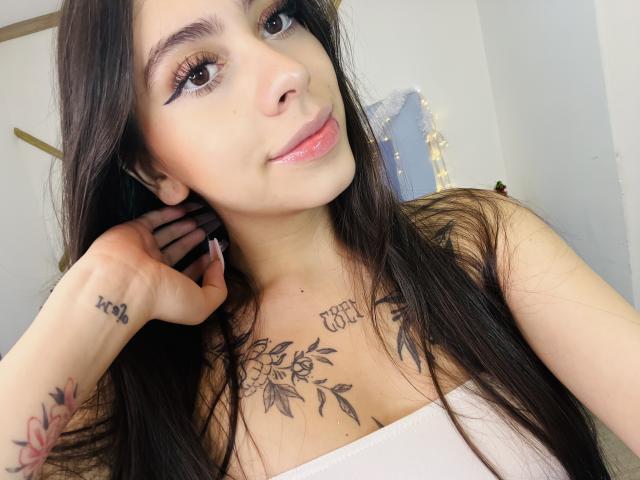 ChanelDeRosee with long dark hair, pink lipstick, and eyeliner, wearing a white top, showing tattoos on chest and forearm, hand near ear, smiling. Boobs visible, soft light.