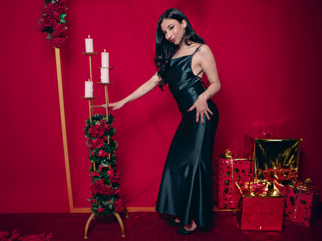 CamilleRios in a black, one-shoulder, satin dress, touching a candle stand with white candles and red roses, next to Christmas gifts on a red carpet.