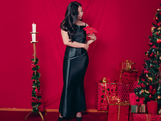 Camille Rios trägt ein schwarzes, glänzendes, eng anliegendes Kleid und hält eine rote Weihnachtssternblume. Sie steht neben einem Kerzenständer und einem Weihnachtsbaum mit Geschenken. Sie hat langes, dunkles Haar..