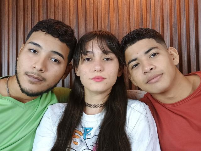 Three BiGroupSexs with cute faces, curly and short hair, wearing green, red, and white shirts, standing closely, wooden background, girl with choker, and all smiling at camera.