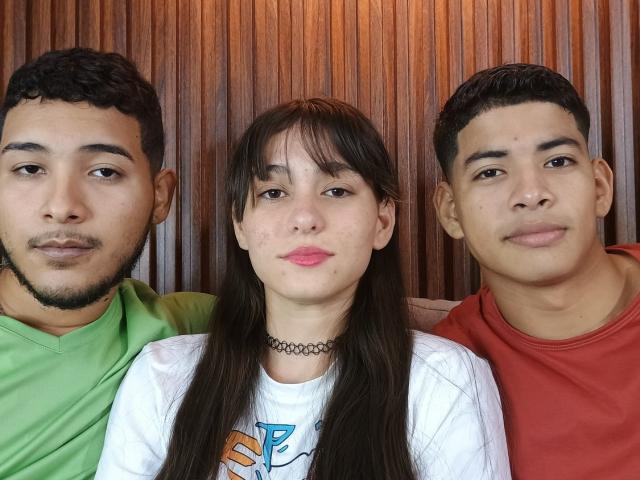 Three BiGroupSex individuals, one with green shirt, one with white shirt and black choker, one with red shirt, standing closely, wooden background, all with short dark hair and neutral expressions.