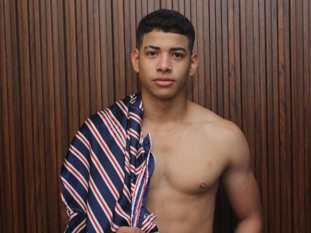 Shirtless BiGroupSex with dark short hair, medium brown skin, and a toned, muscular chest, holding a striped shirt over one shoulder, looking straight at the camera.