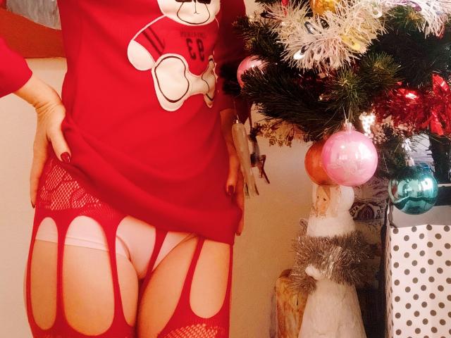 MartaLor draagt een rode jurk met uitsparingen, waardoor haar roze onderbroek en rode garters zichtbaar zijn, en staat naast een kerstboom met versieringen..