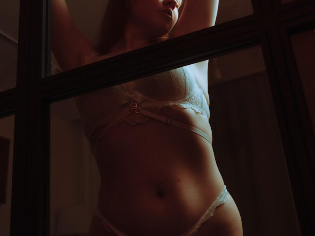 MoonBeauty standing in front of a window, arms raised, wearing white lace bra and panties, soft light highlighting her tits and slightly visible pussy. Dim, moody lighting.