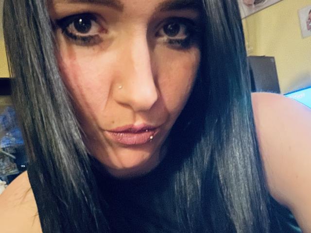 LustLauder has long black hair, brown eyes, and a nose piercing. Her lips are slightly pouted with a lip ring. She's looking directly at the camera with a subtle smirk.