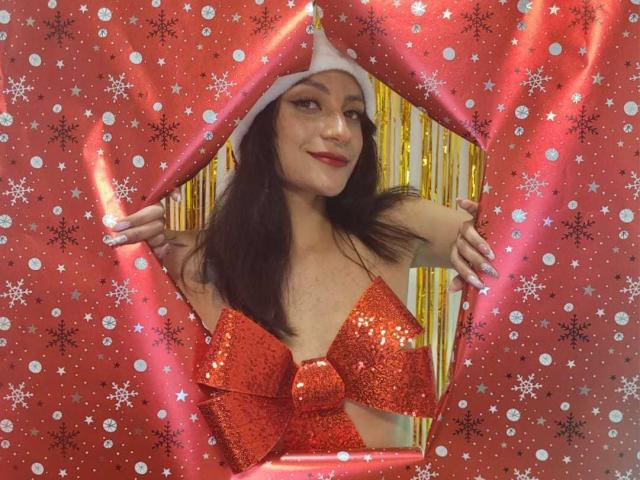 Alizep with dark hair, wearing a white hat and red glittery bikini top, smiling, hands on red, glittery, snowflake-patterned, gift-wrapping paper. Boobs, red glitter, white hat.
