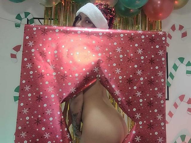 Nude Alizep with a white Santa hat, hiding behind a red, polka-dotted, snowflake-patterned gift, showing their ass and a tattoo on their right hip.