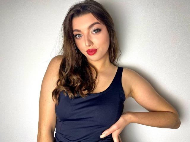 Vailett, met golvend bruin haar, draagt een zwarte tanktop, heeft rode lippenstift en een subtiele glimlach, met haar hand op haar heup, en kijkt met groene ogen in de camera..