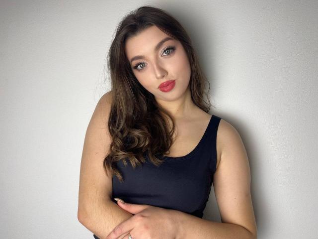 Vailett, con cabello castaño ondulado, vestida con una camiseta negra, con los brazos cruzados, lápiz labial rojo y maquillaje de ojos discreto, mirando directamente a la cámara. Tiene la piel pálida y ojos verdes..