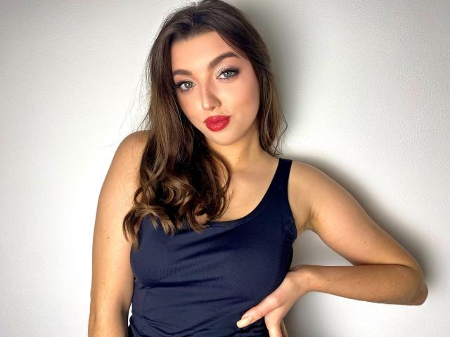 Vailett with wavy brown hair, red lipstick, and dark tank top, hand on hip, looking at camera, showing her tits and ass in a sexy, confident pose.