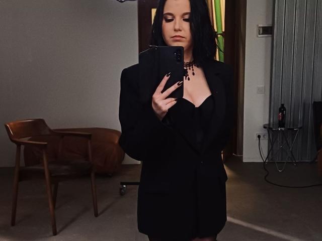 Janel with black hair, wearing a black blazer, taking a mirror selfie with her phone, showing her tits slightly, in a dimly lit room with wooden chairs.