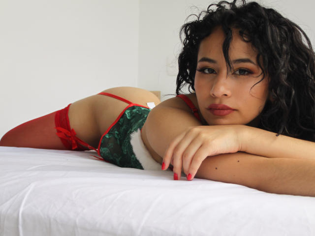 HeilyPeterson lying on a bed, wearing red and green lingerie with white fur trim, red thigh-highs, and red nails, looking at the camera with curly black hair.