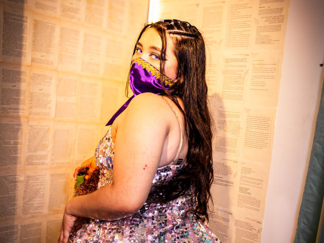 AishaGhulam with long brown braids, purple face mask, and sparkly dress, showing her back, looking over her shoulder, with her ass slightly raised. Her eyes are visible.