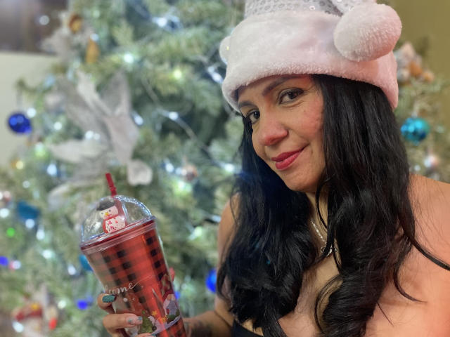 LailaCastillo with black wavy hair, pink fluffy Santa hat, holding red bubble tea, smiling, wearing a necklace, standing in front of a decorated Christmas tree.