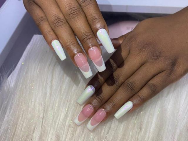 SamyLorenss' hands with long, square-tipped nails, pink and white French tips, and iridescent green tips, resting on a white, glittery surface. Dark brown skin, shiny nails.