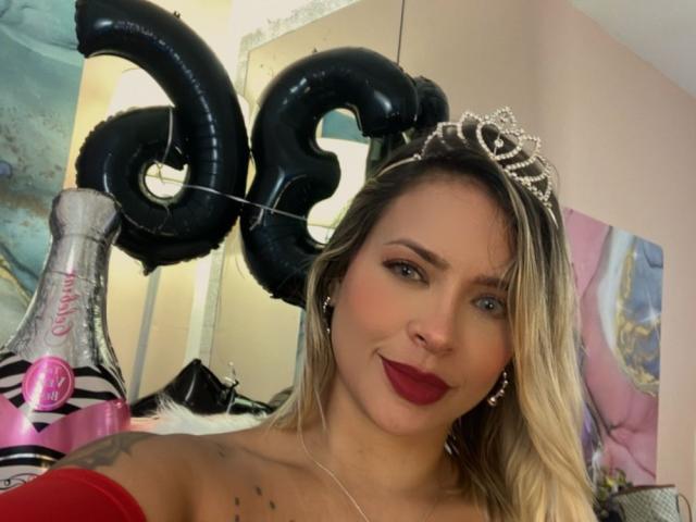 ChantalSex69, con cabello rubio, lápiz labial rojo, una tiara y aretes, vistiendo una blusa roja con un hombro descubierto, sosteniendo una copa de champán, globos negros con el número "28", sonriendo, con tatuajes en el hombro..