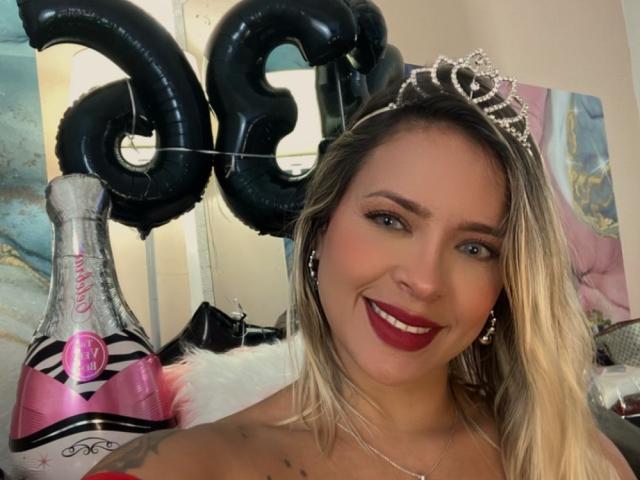 ChantalSex69 with blonde hair, red lipstick, and a tiara, smiling at the camera, holding a bottle and black "28" balloons, showing her bare shoulders and tattoo.