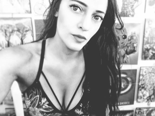 Zwart-wit selfie van JaneRoa met lang donker haar, die een beha met bloemenprint en bandjes draagt, waarbij haar borsten zichtbaar zijn en ze een lichte frons op haar gezicht heeft..