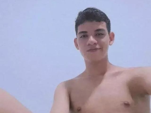 SexyAladinX, shirtless, with short dark hair, light skin, small nipples, and a slight smile, against a clear blue sky. His chest is visible, no other body parts shown.