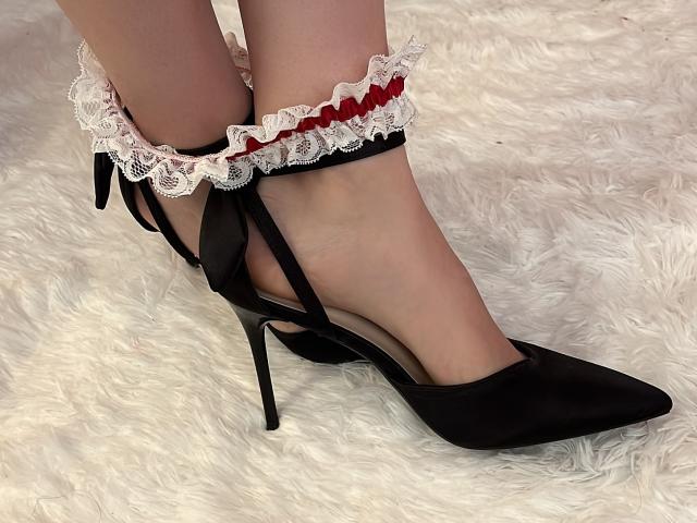 MelanieFrancaise's feet in black high heels with red and white lace ankle straps, sitting on a white fluffy rug. Her toes are pointed, showing off the heels.