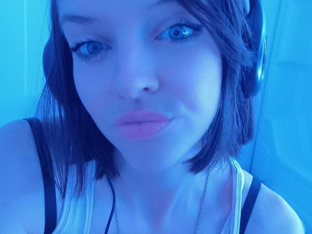 LissaMeow with blue-tinted face, wearing headphones, has short brown hair, blue eyes, and pink lipstick, in a sleeveless top, showing her tits slightly.
