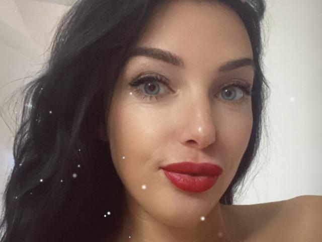 KatieFrenchie's face with dark, wavy hair, blue eyes, and red lipstick, showing a slight smile with small white dots across her skin. Boobs are not visible.
