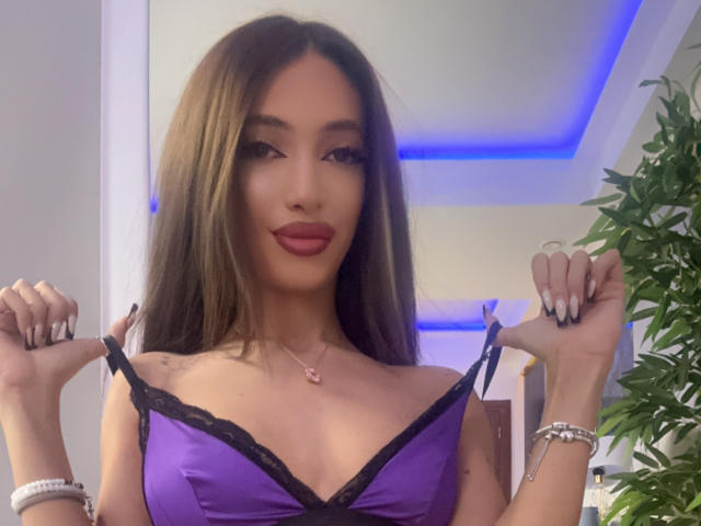 LaylaKrista with long brown hair, wearing a purple satin bra with black lace, holding straps, red lipstick, manicured nails, pink necklace, and bracelets, smiling.