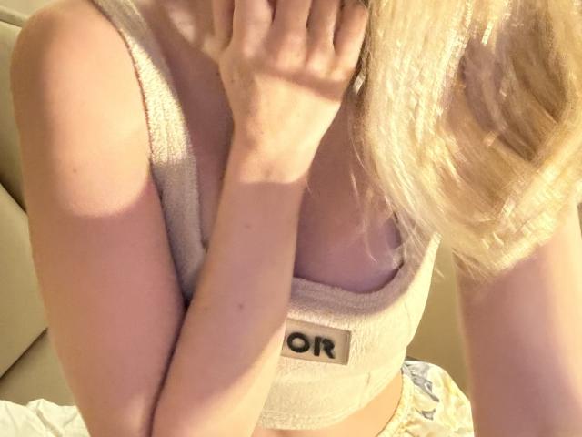 SelinaRose's hand covers her mouth, blonde hair falls over her shoulder, wearing a beige tank top, her small tits slightly visible, sunlight casts soft shadows on her skin.