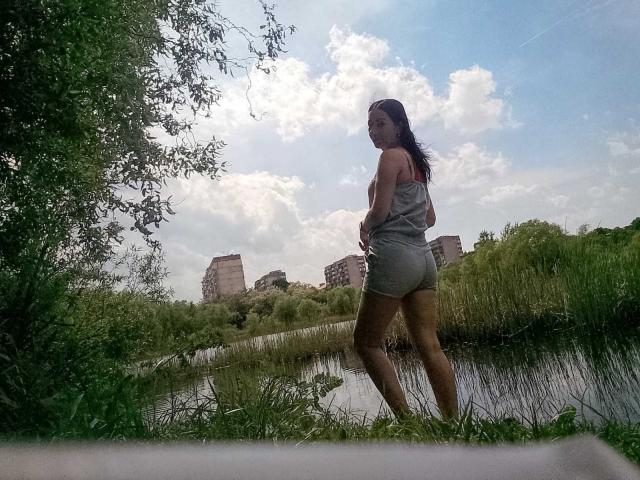 MirandaHott69 with long brown hair, grey tank top, and tight grey shorts, standing by a grassy waterway, showing her ass, looking over her shoulder, sunny day, blue sky.