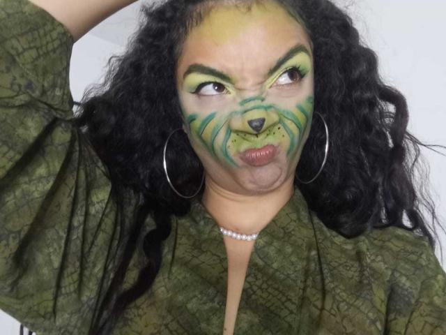 AndromedaZareth with green and yellow face paint resembling a cat, puffed lips, curly black hair, hoop earrings, green textured shirt, and a pearl necklace, making a goofy face.