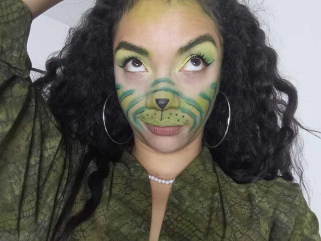 AndromedaZareth with green and yellow cat face makeup, curly black hair, large hoop earrings, green patterned shirt, pearl necklace, and serious expression. Boobs visible under shirt.