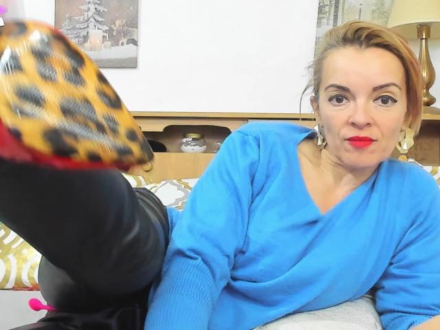 KateRouge with red lipstick, blue sweater, and black pants, raising her leopard-print shoe, looking at the camera with a playful expression. Her tits are not visible.