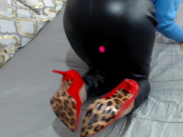 KateRouge's ass in tight black latex, wearing leopard print and red high heels, kneeling on a gray bed with blue sleeves visible.